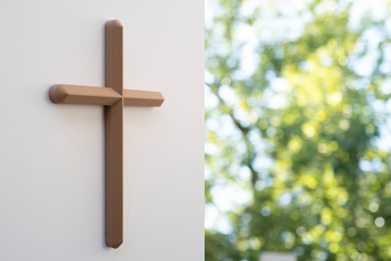 May include: A brown wooden cross mounted on a white wall. The cross is in focus, while the background is blurred and shows a green leafy tree.