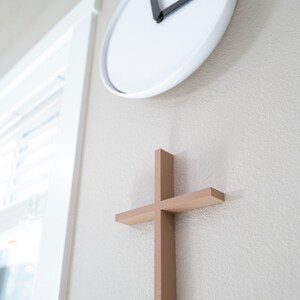 Peut inclure: Une horloge blanche avec des aiguilles noires est mont&eacute;e sur un mur beige clair, avec une croix en bois mont&eacute;e en dessous.
