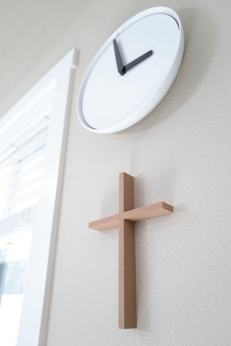 Peut inclure: Une horloge blanche avec des aiguilles noires est mont&eacute;e sur un mur beige clair, avec une croix en bois mont&eacute;e en dessous.