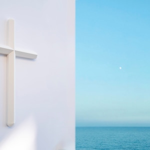 May include: A white cross is mounted on a white wall with a view of a blue ocean and a sliver of a white moon in the sky.