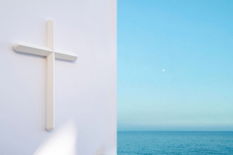 May include: A white cross is mounted on a white wall with a view of a blue ocean and a sliver of a white moon in the sky.