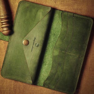 Op de afbeelding: Open, olijfgroene leren portemonnee met een drukknoopsluiting. De portemonnee is open en toont binnenvakken en een flap met de tekst "Langlous Leather" en een logo. Op een houten oppervlak.
