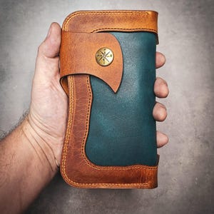 Può includere: Un portafoglio in pelle fatto a mano nei toni del marrone e del verde acqua. Il portafoglio presenta un design a bussola su una chiusura con bottone in ottone. I bordi sono cuciti con un filo a contrasto e il portafoglio è tenuto in una mano.
