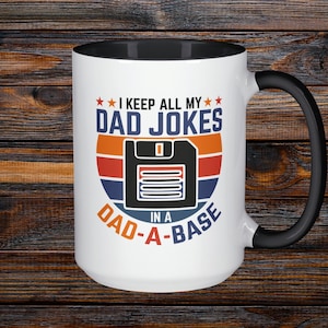 May include: White ceramic mug with a black handle and interior. The mug features the text "I KEEP ALL MY DAD JOKES in a DAD-A-BASE" with a retro design of a floppy disk. The mug is on a dark wood surface.