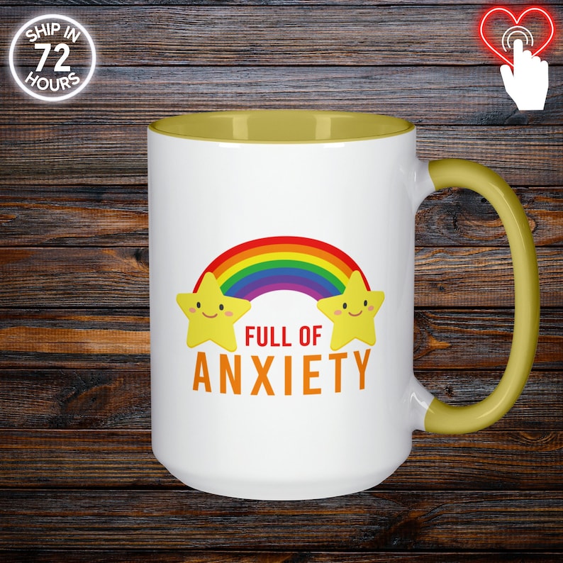 May include: White ceramic mug with a yellow interior and handle. The mug features a rainbow and two smiling yellow stars. The words "FULL OF ANXIETY" are printed in orange. The mug is on a dark wood background.