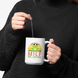May include: White ceramic mug featuring a cartoon frog design with a sunset background. The mug has the text "MILF MAN, I LOVE FROGS" in orange and green. The mug is held by a person wearing a black sweater.