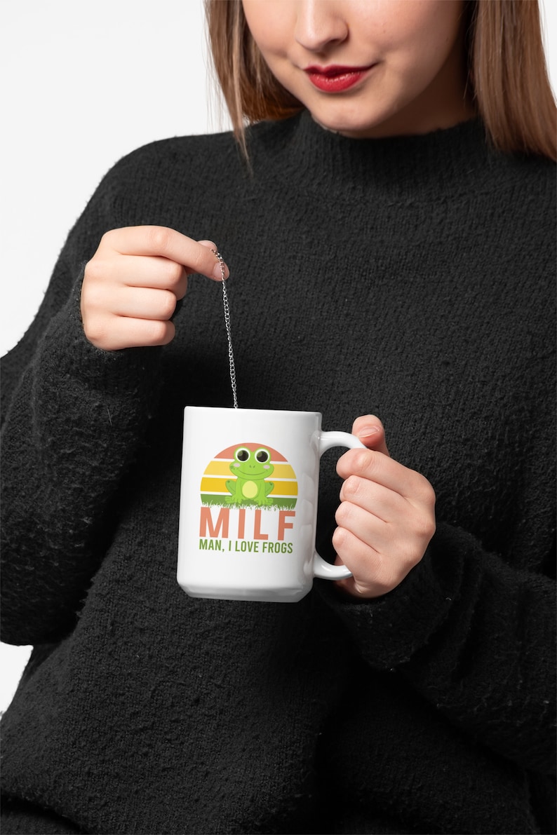 May include: White ceramic mug featuring a cartoon frog design with a sunset background. The mug has the text "MILF MAN, I LOVE FROGS" in orange and green. The mug is held by a person wearing a black sweater.