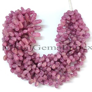 May include: A strand of pink gemstone beads. The beads are oval-shaped and have a polished surface. The color is a medium pink with some variation in tone. The beads are strung together on a white cord. The text "Mittal Gems Impex" is visible.