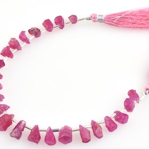 Op de afbeelding: Een streng roze robijn edelsteen kralen, elke kraal heeft een ruwe, onregelmatige vorm. De kralen zijn geregen op zilverdraad en hebben aan elk uiteinde roze kwastjes.
