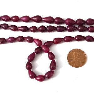 May include: Strands of deep red teardrop-shaped beads. The beads are arranged in rows and a circle. A penny is included for size reference. The beads have a glossy finish and are likely for jewelry making.