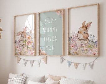 May include: Three framed prints with a light blue background. The center print has the text "Some Bunny Loves You". The left and right prints feature watercolor illustrations of bunnies with flowers. The prints are hanging on a white wall above a wooden bench with storage bins.