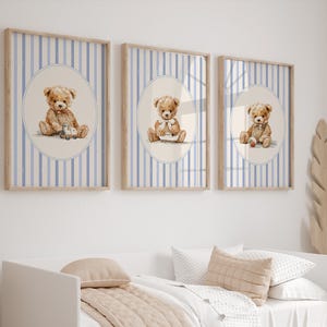 May include: Three framed prints featuring watercolor illustrations of teddy bears. Each bear is depicted in a different pose, holding a toy or wearing a bow. The background has blue and white vertical stripes, and the frames are light brown.
