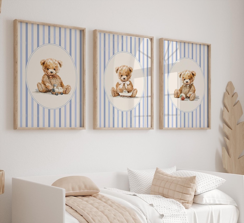 May include: Three framed prints featuring watercolor illustrations of teddy bears. Each bear is depicted in a different pose, holding a toy or wearing a bow. The background has blue and white vertical stripes, and the frames are light brown.