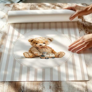 May include: A rolled-up print featuring a watercolor illustration of a teddy bear with a toy train. The bear is light brown with a pink bow tie, set against a striped background in beige and white. The print is being unrolled on a wooden surface.