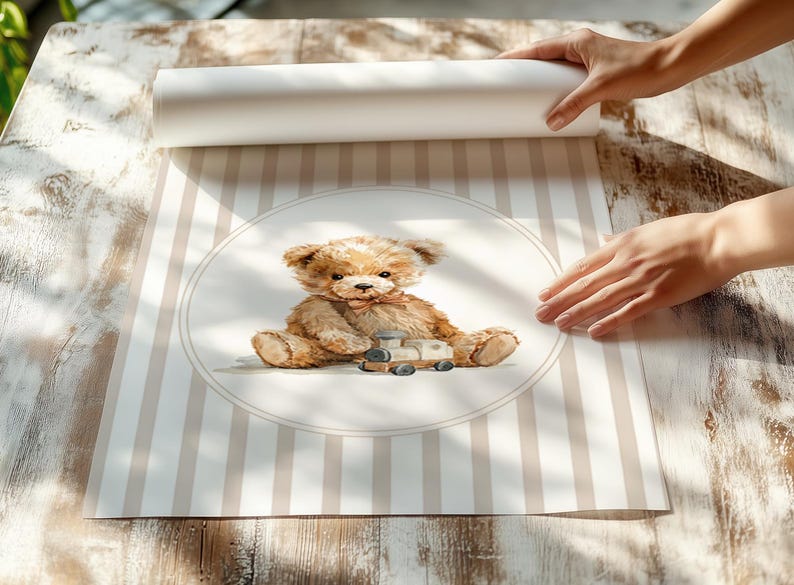 May include: A rolled-up print featuring a watercolor illustration of a teddy bear with a toy train. The bear is light brown with a pink bow tie, set against a striped background in beige and white. The print is being unrolled on a wooden surface.