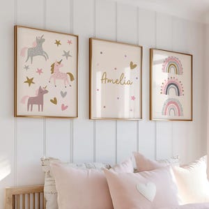 K&ouml;nnte beinhalten: Drei gerahmte Wandkunstwerke mit goldenen Rahmen. Eines zeigt Einh&ouml;rner, Sterne und Herzen in Rosa, Grau und Gold. Ein anderes hat den Namen "Amelia" mit Sternen und einem Herzen. Das dritte zeigt Regenbogen-Designs.