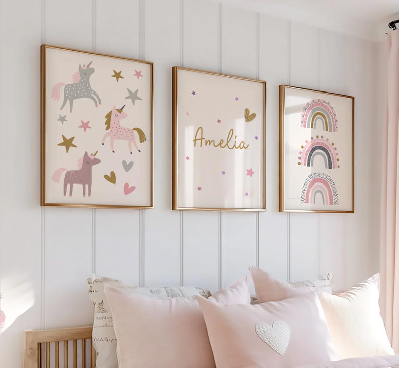 K&ouml;nnte beinhalten: Drei gerahmte Wandkunstwerke mit goldenen Rahmen. Eines zeigt Einh&ouml;rner, Sterne und Herzen in Rosa, Grau und Gold. Ein anderes hat den Namen "Amelia" mit Sternen und einem Herzen. Das dritte zeigt Regenbogen-Designs.