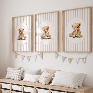 May include: Three framed prints featuring watercolor illustrations of teddy bears. Each bear is depicted in a different pose, set against a striped background within an oval frame. The frames are light wood, and the overall theme is a nursery or children's room.