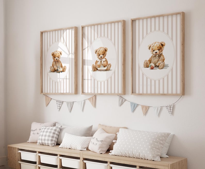 May include: Three framed prints featuring watercolor illustrations of teddy bears. Each bear is depicted in a different pose, set against a striped background within an oval frame. The frames are light wood, and the overall theme is a nursery or children's room.