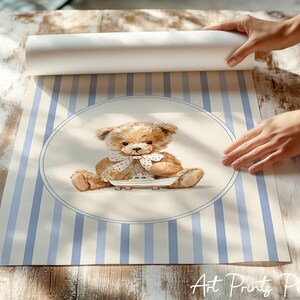 May include: A watercolor print featuring a teddy bear with a white bow tie, sitting on a small boat. The background has blue and white vertical stripes. The print is being unrolled on a wooden surface.