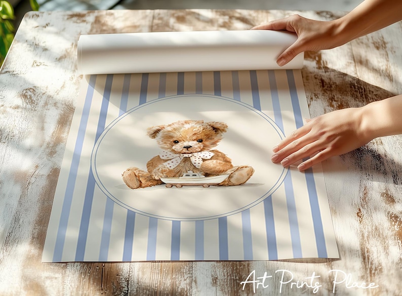 May include: A watercolor print featuring a teddy bear with a white bow tie, sitting on a small boat. The background has blue and white vertical stripes. The print is being unrolled on a wooden surface.