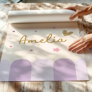 May include: A personalized poster with the name "Amelia" in gold script, adorned with pink and purple stars, and a gold heart. The bottom features a row of purple arches. The poster is being unrolled on a wooden surface.