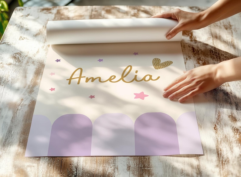 May include: A personalized poster with the name "Amelia" in gold script, adorned with pink and purple stars, and a gold heart. The bottom features a row of purple arches. The poster is being unrolled on a wooden surface.