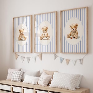 May include: Three framed prints featuring watercolor teddy bears. Each print has a light blue and white striped background with an oval containing a teddy bear. The frames are light brown. A garland of small flags hangs below the prints.