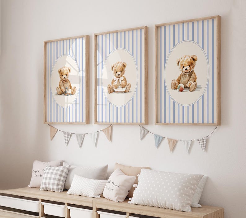 May include: Three framed prints featuring watercolor teddy bears. Each print has a light blue and white striped background with an oval containing a teddy bear. The frames are light brown. A garland of small flags hangs below the prints.