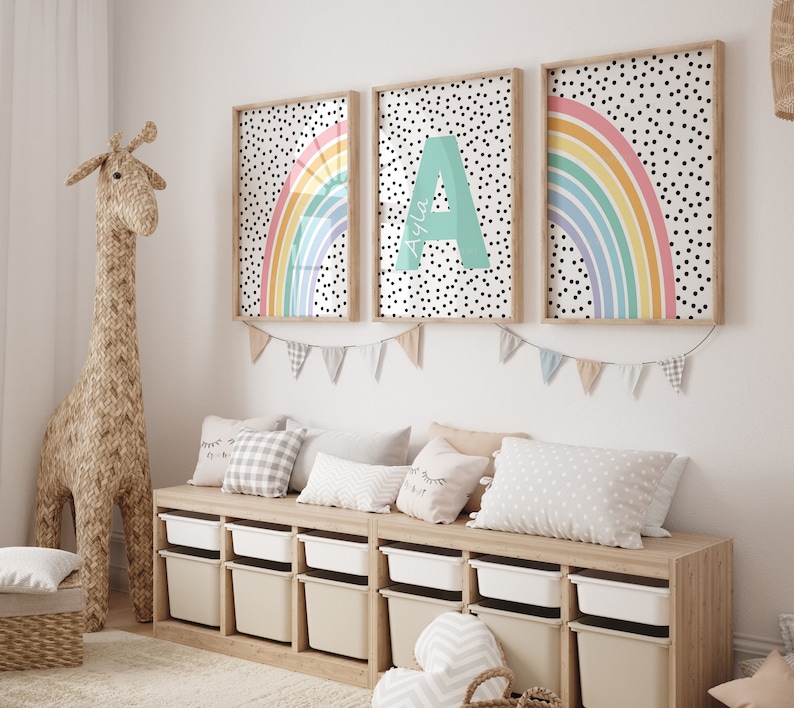 May include: Three framed prints featuring a rainbow and polka dot design. The centre print displays the letter A with the name Ayla written below. The prints are hung on a white wall above a wooden storage bench with white storage bins.
