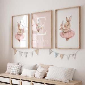 May include: Three framed prints with a pink background. The center print features a large letter E with a floral design and the name Evelyn. The prints on either side feature a bunny wearing a pink tutu and holding flowers.