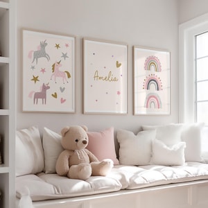 K&ouml;nnte beinhalten: Drei gerahmte Kunstdrucke mit hellen Holzrahmen. Die Drucke zeigen Einh&ouml;rner, den Namen "Amelia" und Regenbogen. Die Farbpalette umfasst Rosa, Gold, Grau und Wei&szlig;. Ein Teddyb&auml;r und Kissen befinden sich auf einer Fensterbank unter der Kunst.
