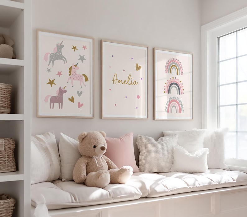 K&ouml;nnte beinhalten: Drei gerahmte Kunstdrucke mit hellen Holzrahmen. Die Drucke zeigen Einh&ouml;rner, den Namen "Amelia" und Regenbogen. Die Farbpalette umfasst Rosa, Gold, Grau und Wei&szlig;. Ein Teddyb&auml;r und Kissen befinden sich auf einer Fensterbank unter der Kunst.