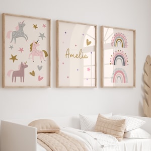 K&ouml;nnte beinhalten: Drei gerahmte Wandkunstdrucke mit hellem Holzrahmen. Die Drucke zeigen Einhorn-Illustrationen, den Namen "Amelia" und Regenbogen-Designs in sanften Rosa-, Gold- und Graut&ouml;nen. Die Kunstwerke werden &uuml;ber einem Bett mit neutralfarbener Bettw&auml;sche ausgestellt.