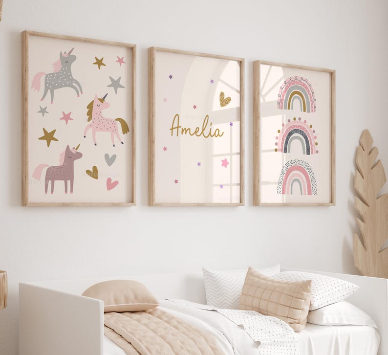 K&ouml;nnte beinhalten: Drei gerahmte Wandkunstdrucke mit hellem Holzrahmen. Die Drucke zeigen Einhorn-Illustrationen, den Namen "Amelia" und Regenbogen-Designs in sanften Rosa-, Gold- und Graut&ouml;nen. Die Kunstwerke werden &uuml;ber einem Bett mit neutralfarbener Bettw&auml;sche ausgestellt.