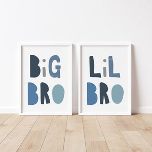 May include: Two framed prints with the words "BIG BRO" and "Lil BRO" in shades of blue. The prints are in white frames and are displayed on a light wood floor. The wall behind the prints is white.