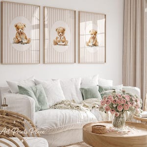 May include: Three framed art prints featuring watercolor teddy bears. Each print has a beige striped background with an oval containing a teddy bear illustration. The bears are light brown and have different accessories.