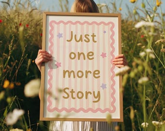May include: A framed print with the words "Just one more Story" in gold lettering. The print has a pink and white striped background with pink scalloped edges and star accents. The frame is a light brown colour.