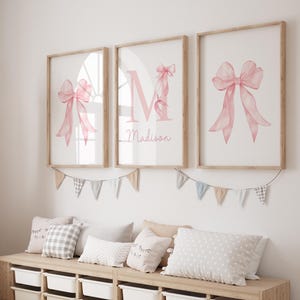 Puede incluir: Tres impresiones enmarcadas con lazos rosas de acuarela y un marco de madera. La impresión central presenta la letra "M" y el nombre "Madison". Una guirnalda de pequeñas banderas cuelga debajo. Debajo hay almohadas y un banco de almacenamiento.
