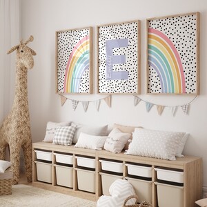 May include: Three framed prints featuring a rainbow and polka dot design. The centre print displays the letter "E" in a pale purple hue. The prints are hung on a white wall above a wooden storage bench with white containers.