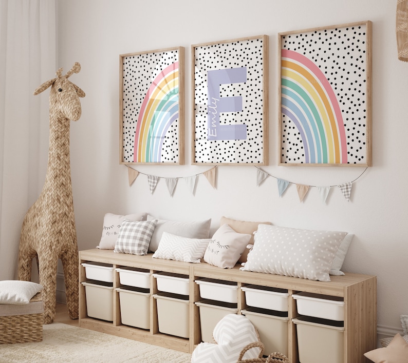 May include: Three framed prints featuring a rainbow and polka dot design. The centre print displays the letter "E" in a pale purple hue. The prints are hung on a white wall above a wooden storage bench with white containers.