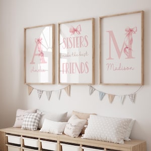 Puede incluir: Tres impresiones artísticas enmarcadas con marcos de madera clara. Las impresiones presentan letras y lazos rosas, con el texto "Sisters make the best friends". Debajo, los nombres "Amelia" y "Madison" están escritos en cursiva. Una guirnalda de pequeñas banderas cuelga debajo.