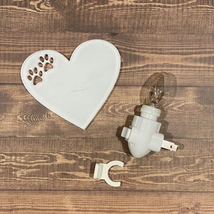 May include: White ceramic heart-shaped nightlight with paw print cutouts. The nightlight has a white plug-in base with a clear light bulb.