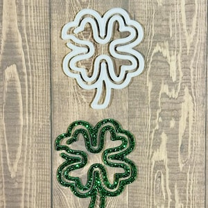 May include: Two four-leaf clover decorations. One is white, the other is green with glitter. The clovers are on a wooden background. Ideal for St. Patrick's Day.