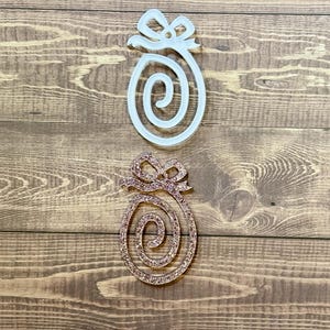 May include: Two Christmas ornaments shaped like a gift with a bow. The top ornament is white and the bottom ornament is a sparkly rose gold colour.