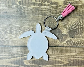 May include: A light blue sea turtle keychain with a pink tassel. The keychain is made of a translucent material and has a silver-colored metal ring. The tassel is a bright pink color and is attached to the keychain with a silver-colored metal cap.