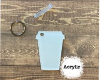 May include: A light blue acrylic coffee cup-shaped keychain with a gold key ring, a clear straw, and the word "Acrylic" on a white circle, all on a wooden background.