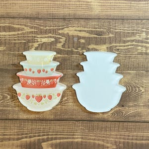 May include: A stack of vintage Pyrex nesting bowls with a strawberry pattern. The bowls are in shades of red, yellow, and white. The bowls are arranged on a wooden surface.