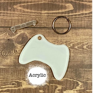 May include: A white acrylic blank video game controller keychain with a silver key ring. The keychain is on a brown wooden surface.