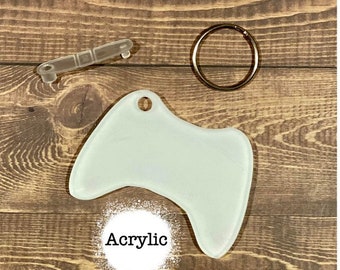 May include: A white acrylic blank video game controller keychain with a silver key ring. The keychain is on a brown wooden surface.